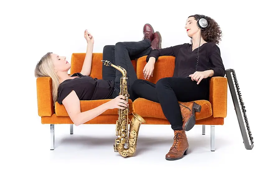 Pressefoto des Duos Breuer & Hermenau mit zwei Musikerinnen vor weißem Hintergrund auf einem orangefarbenen Sofa, eine mit Saxofon liegend auf dem Sofa, die andere mit Kopfhörern sitzend, daneben ein Akkordeon.