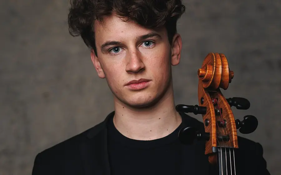 Der Cellist Lenz Defregger in schwarzem Pullover mit Instrument vor verschwommenem dunkelgrauem Hintergrund
