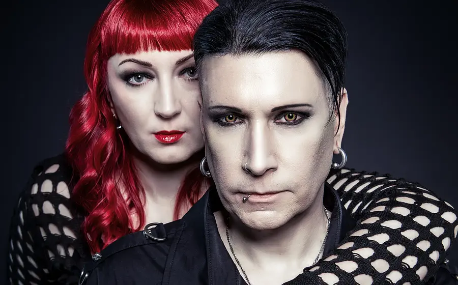 Bandfoto von Blutengel mit zwei Personen in schwarzer Gothic-Kleidung vor dunklem Hintergrund, eine Person mit rotem Haar steht hinter der anderen und legt einen Arm über deren Schulter.