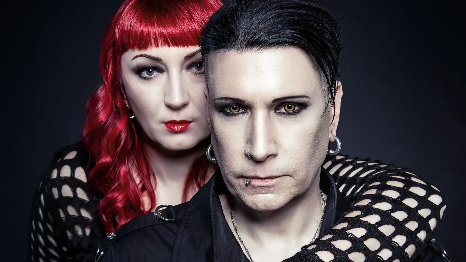 Bandfoto von Blutengel mit zwei Personen in schwarzer Gothic-Kleidung vor dunklem Hintergrund, eine Person mit rotem Haar steht hinter der anderen und legt einen Arm über deren Schulter.