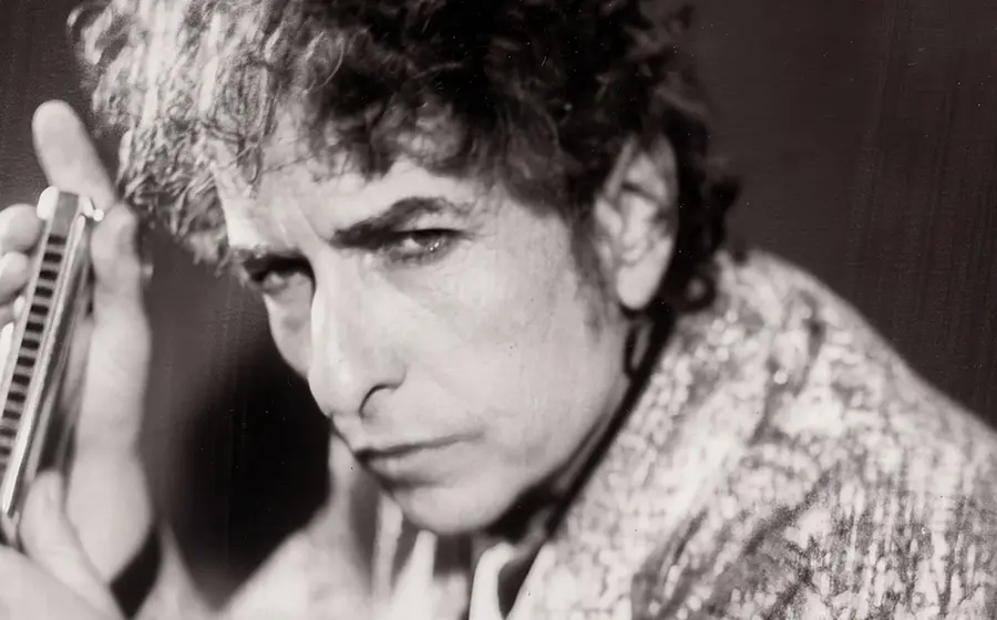 Schwarz-weiss Portrait von Bob Dylan, der seitlich in die Kamer blickt und eine Mundharmonika in den Händen hält, aufgenommen im Abassador Hotel im Jahr 1999