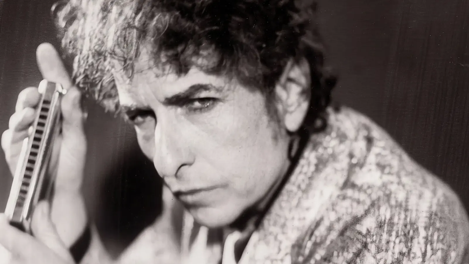 Schwarz-weiss Portrait von Bob Dylan, der seitlich in die Kamer blickt und eine Mundharmonika in den Händen hält, aufgenommen im Abassador Hotel im Jahr 1999