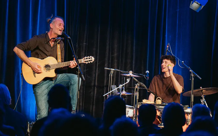 Zwei Musiker treten vor Publikum auf einer Bühne auf, eine Person spielt Akustikgitarre und singt ins Mikrofon, während die andere Person am Schlagzeug sitzt und ein Percussioninstrument hält