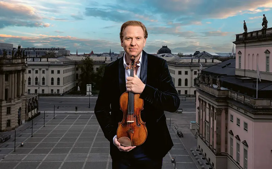 Daniel Hope in dunklem Anzug hält eine Violine vor klassischer Stadtarchitektur mit blau‑orangefarbenem Himmel und großem Stadtplatz im Hintergrund