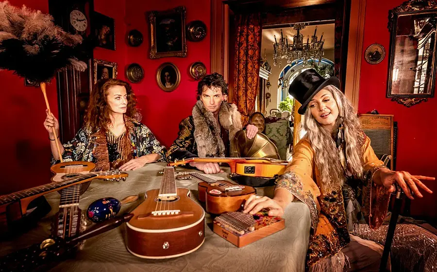 Hippie Kammerorchester: Drei Musikerinnen und Musiker sitzen in einem farbenreichen Raum im Vintage-Stil, umgeben von verschiedenen Instrumenten wie Gitarren und Ukulelen, warme Lichtstimmung.