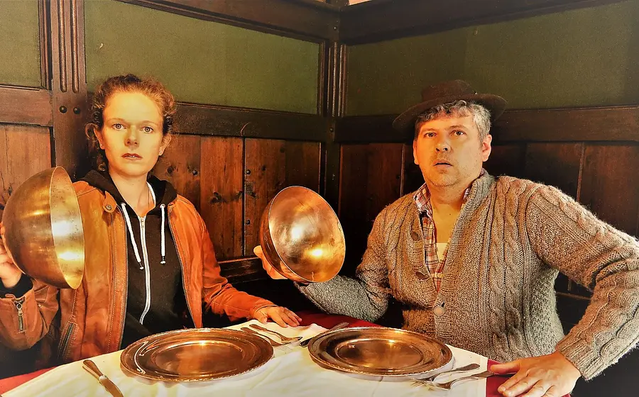 Zwei Personen an einem gedeckten Tisch in holzvertäfeltem Gastraum, beide halten große Metallschüsseln in den Händen und sitzen vor Tellern und Besteck mit überraschter Mimik zur Veranstaltung das Knödelkomplott