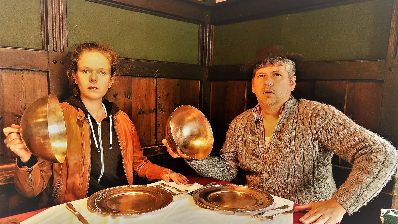 Zwei Personen an einem gedeckten Tisch in holzvertäfeltem Gastraum, beide halten große Metallschüsseln in den Händen und sitzen vor Tellern und Besteck mit überraschter Mimik zur Veranstaltung das Knödelkomplott