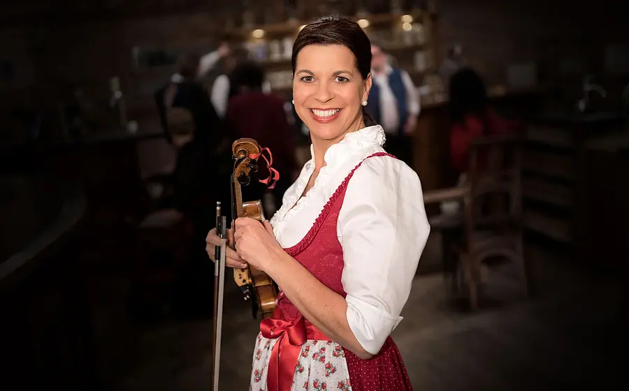 Person in traditioneller Tracht mit rotem Dirndl steht in einem Wirtshaus und hält eine Violine, im Hintergrund unscharf Gäste und Holzmobiliar in gemütlicher Atmosphäre