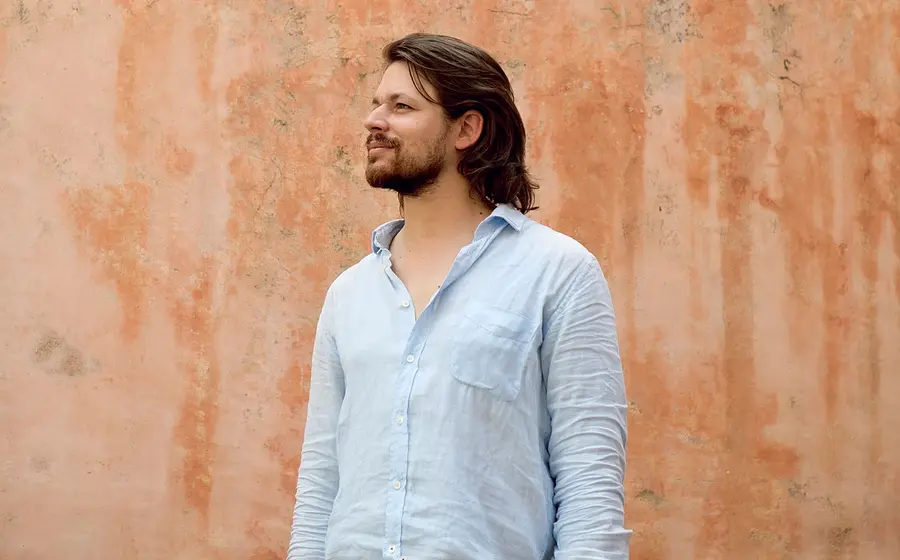 David Fray steht vor einer strukturierten Wand und trägt ein hellblaues Hemd, der Blick ist zur Seite gerichtet.