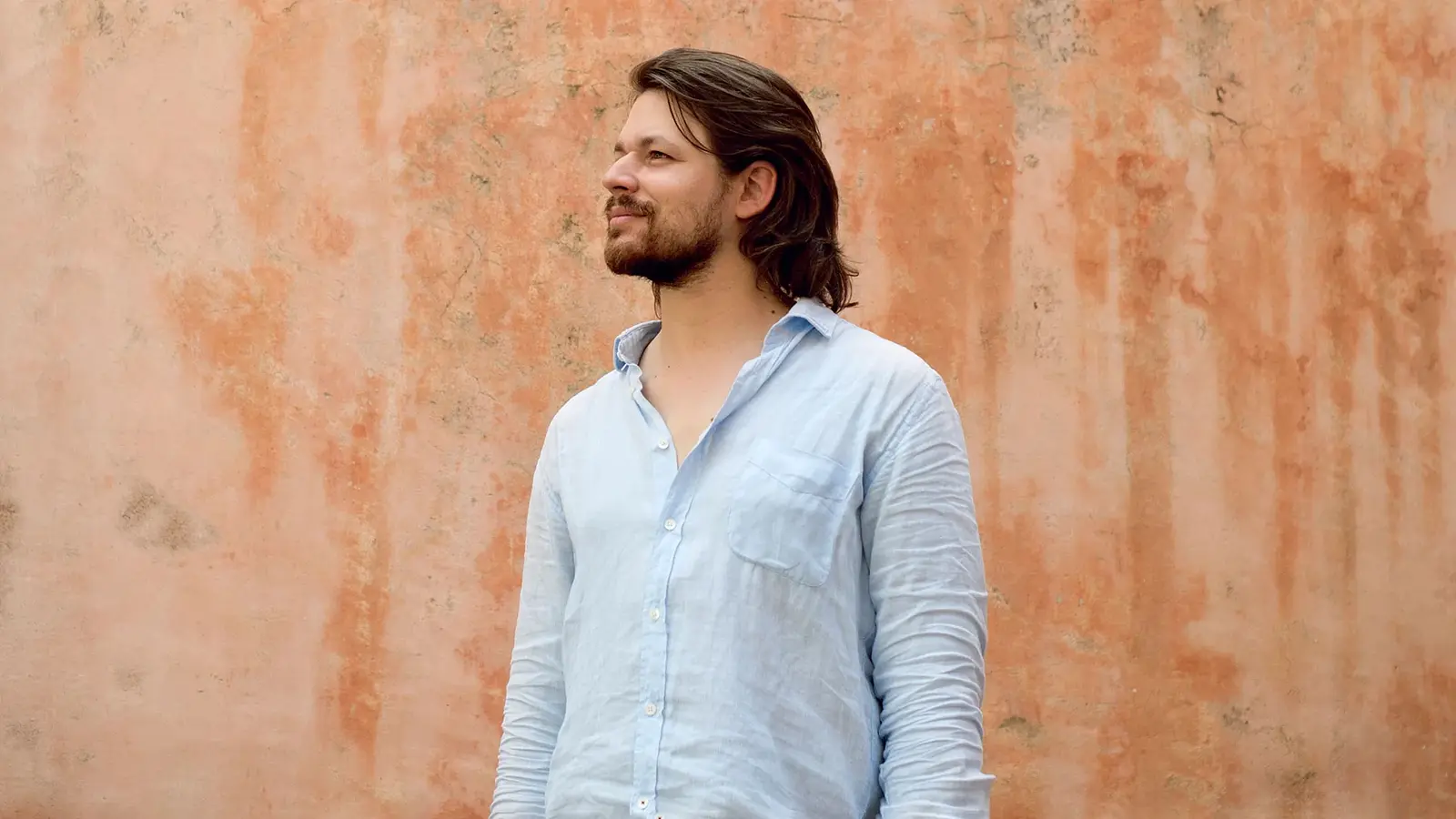 David Fray steht vor einer strukturierten Wand und trägt ein hellblaues Hemd, der Blick ist zur Seite gerichtet.