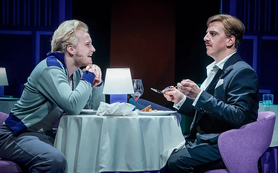 Theaterszene in einem Restaurant. Zwei Darstellende sitzen sich an einem gedeckten Tisch mit Tischlampe, Weinglas und Dessert gegenüber, während eine Person im Anzug mit Besteck in der Hand spricht und die andere sich über den Tisch lehnt.