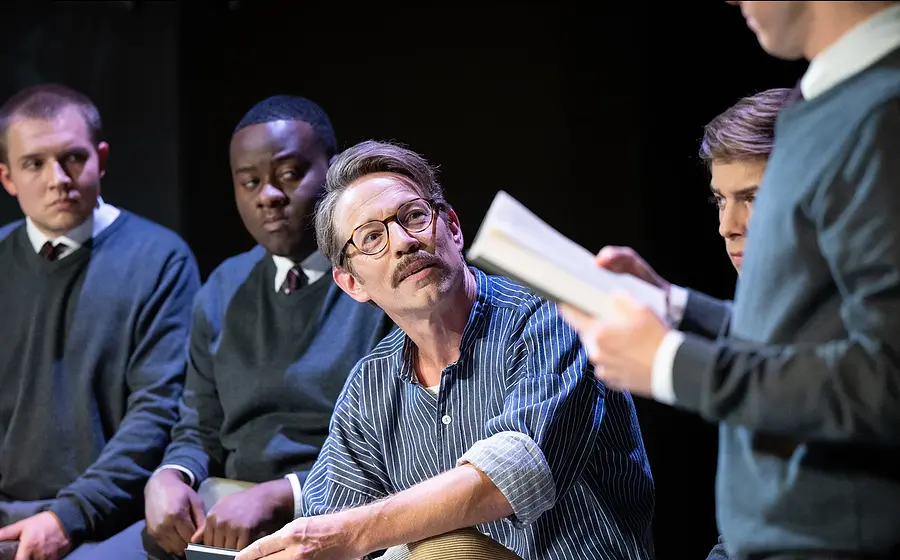 Schauspielszene mit mehreren Darstellern in Schuluniformen und einer sitzenden Person mit Brille, die einem Vorlesenden auf der Bühne zuhört
