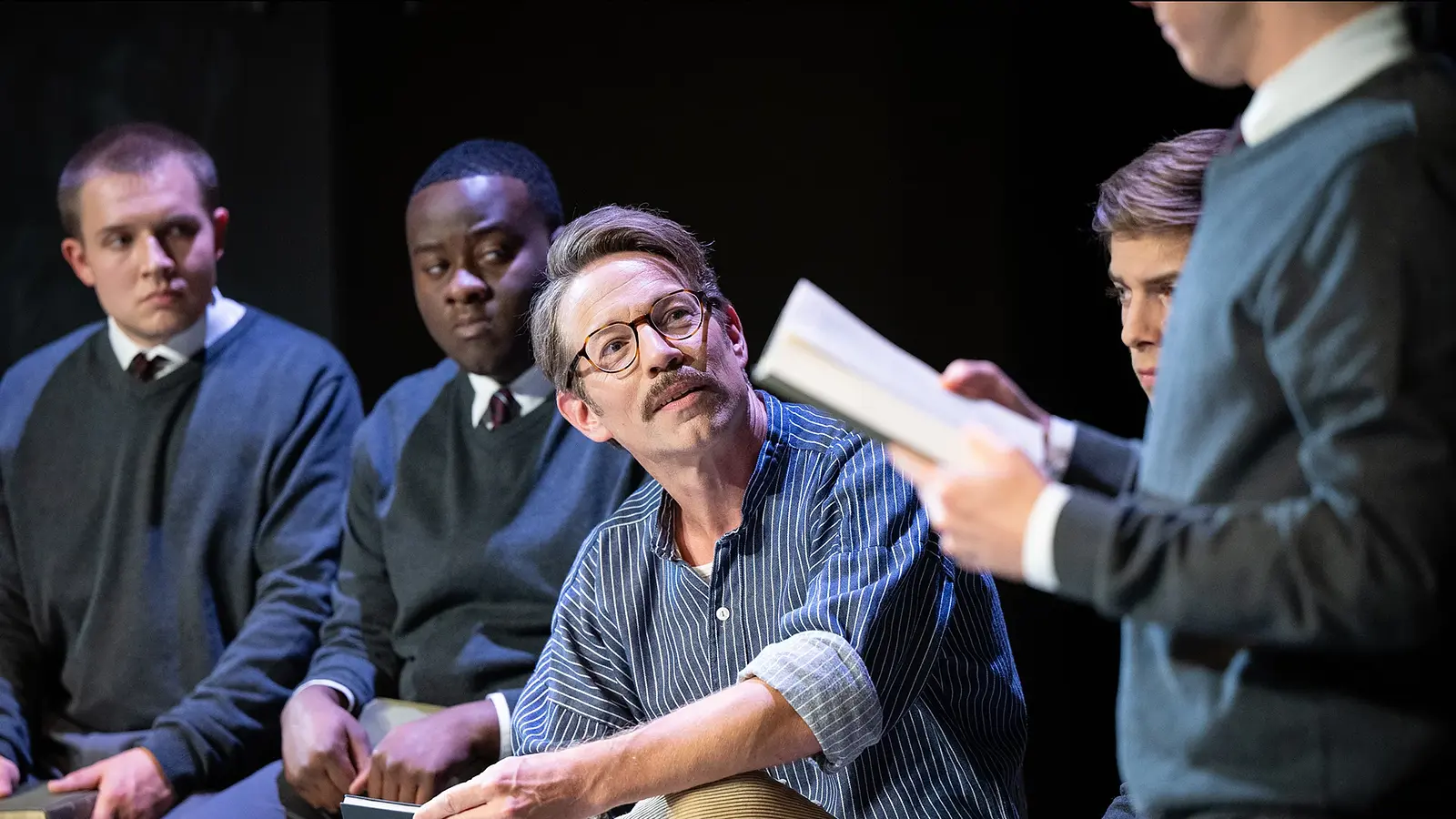 Schauspielszene mit mehreren Darstellern in Schuluniformen und einer sitzenden Person mit Brille, die einem Vorlesenden auf der Bühne zuhört