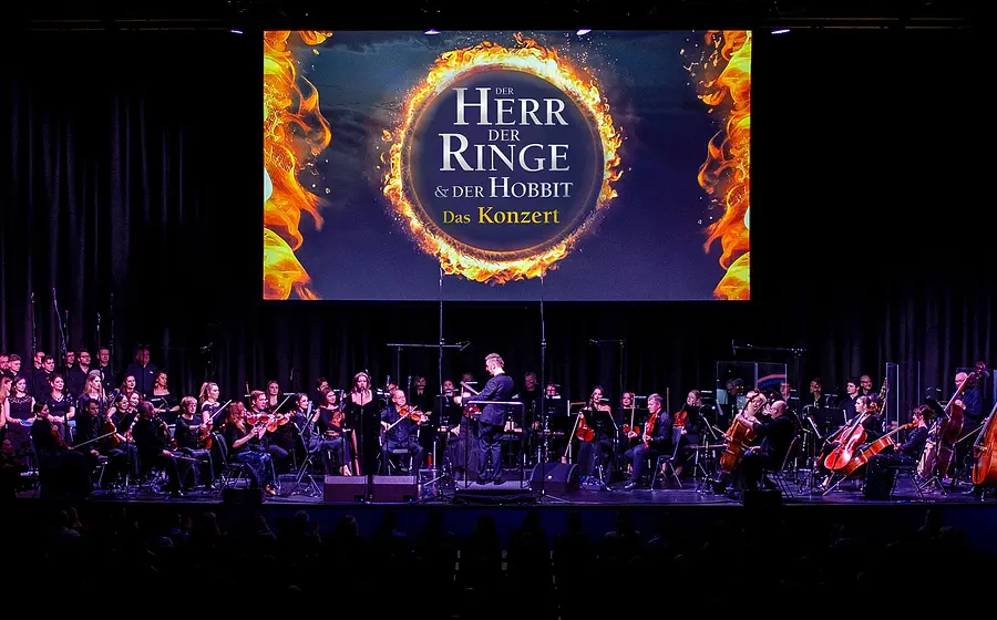 Bühnenfoto zum Konzert „Der Herr der Ringe & Der Hobbit – Das Konzert“ mit großem Orchester und Chor auf einer Konzertbühne, Dirigent in der Mitte, darüber eine große Leinwand mit dem Schriftzug „Der Herr der Ringe & Der Hobbit – Das Konzert“ und einem brennenden Ring.