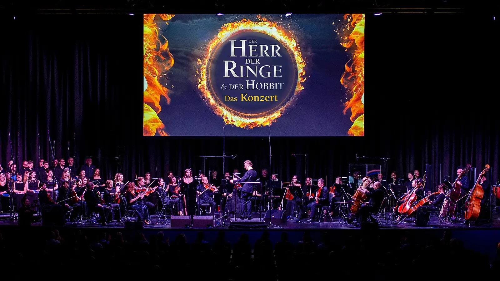 Bühnenfoto zum Konzert „Der Herr der Ringe & Der Hobbit – Das Konzert“ mit großem Orchester und Chor auf einer Konzertbühne, Dirigent in der Mitte, darüber eine große Leinwand mit dem Schriftzug „Der Herr der Ringe & Der Hobbit – Das Konzert“ und einem brennenden Ring.