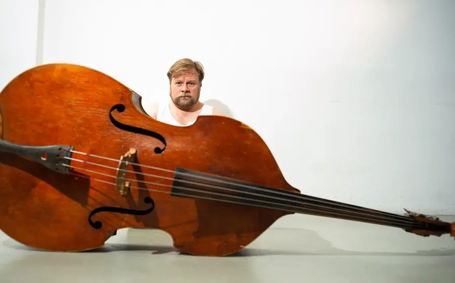 Eine Person sitzt hinter einem auf dem Boden liegenden Kontrabass vor weißem Hintergrund