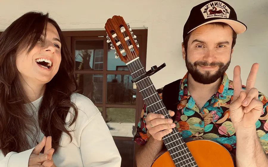 Das Duo mit Akustikgitarre vor einem Gebäude. Eine Person hält die Gitarre und zeigt ein Friedenszeichen, während die andere Person daneben lacht