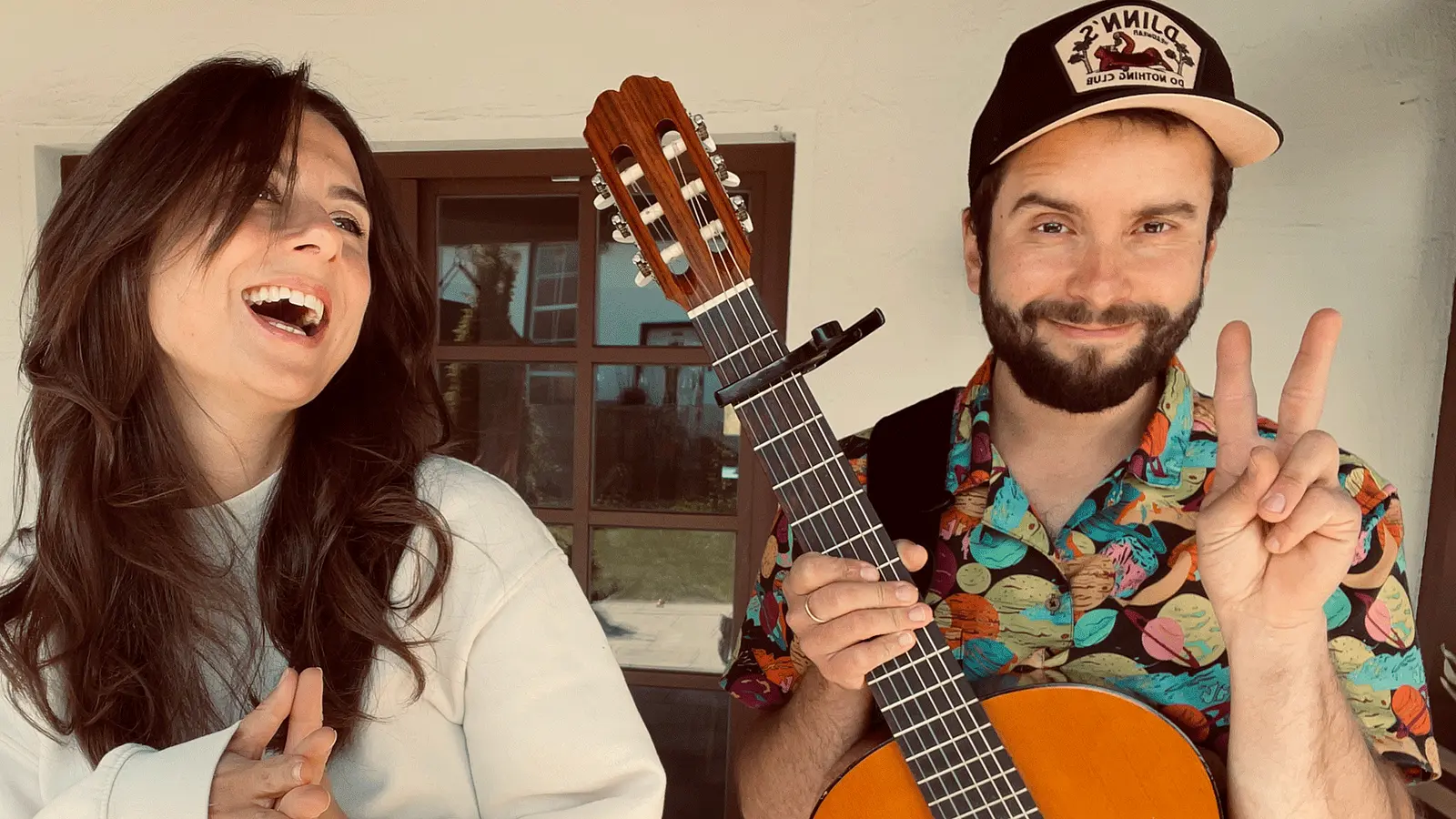 Das Duo mit Akustikgitarre vor einem Gebäude. Eine Person hält die Gitarre und zeigt ein Friedenszeichen, während die andere Person daneben lacht