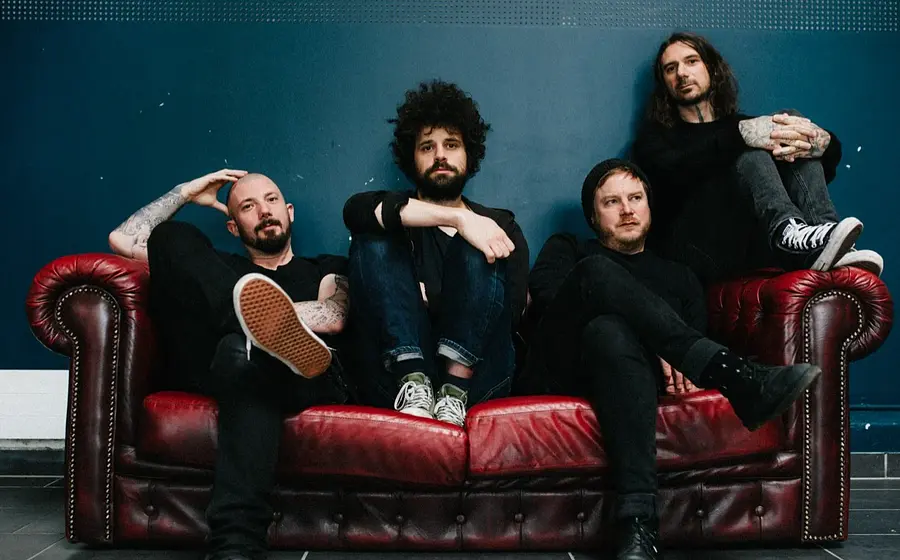 Die Band Coilguns sitzt auf einem antik wirkenden roten Ledersofa mit dunkelblauem Hintergrund