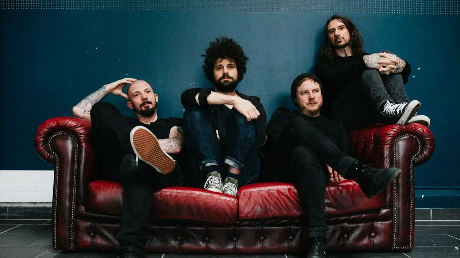 Die Band Coilguns sitzt auf einem antik wirkenden roten Ledersofa mit dunkelblauem Hintergrund