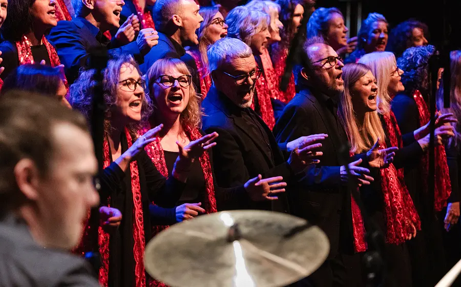 Großer Gospelchor in schwarzer Kleidung mit roten Schals singt auf einer Bühne unter blau roter Beleuchtung. Im Vordergrund ist unscharf ein Schlagzeug zu sehen, während die Chormitglieder in mehreren Reihen auftreten.