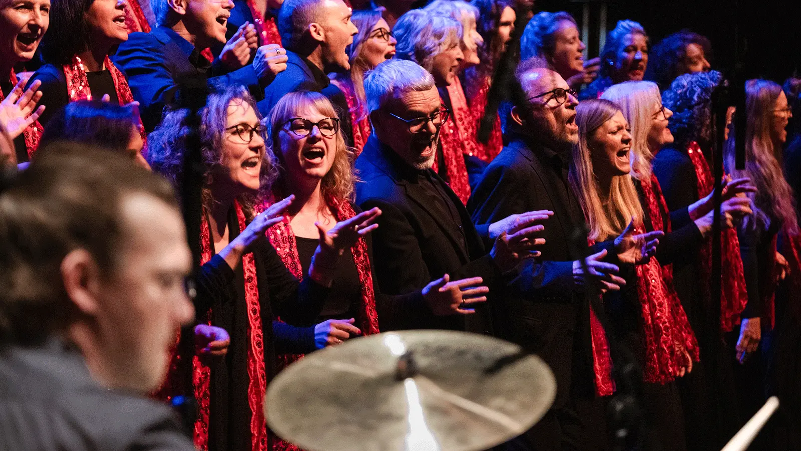 Großer Gospelchor in schwarzer Kleidung mit roten Schals singt auf einer Bühne unter blau roter Beleuchtung. Im Vordergrund ist unscharf ein Schlagzeug zu sehen, während die Chormitglieder in mehreren Reihen auftreten.