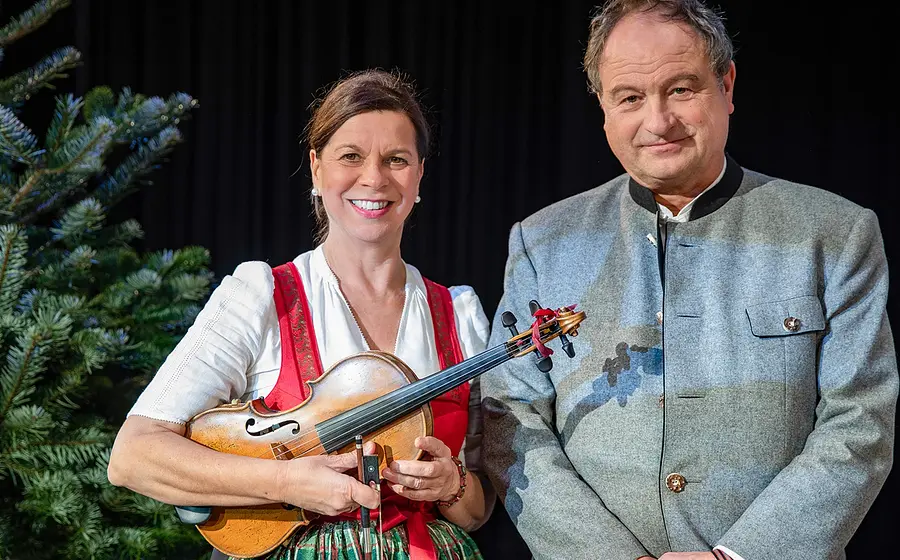 Zwei Personen neben einem Weihnachtsbaum vor schwarzem Hintergrund, eine Person in roter Tracht hält eine Violine in den Händen, die andere trägt eine graue Trachtenjacke