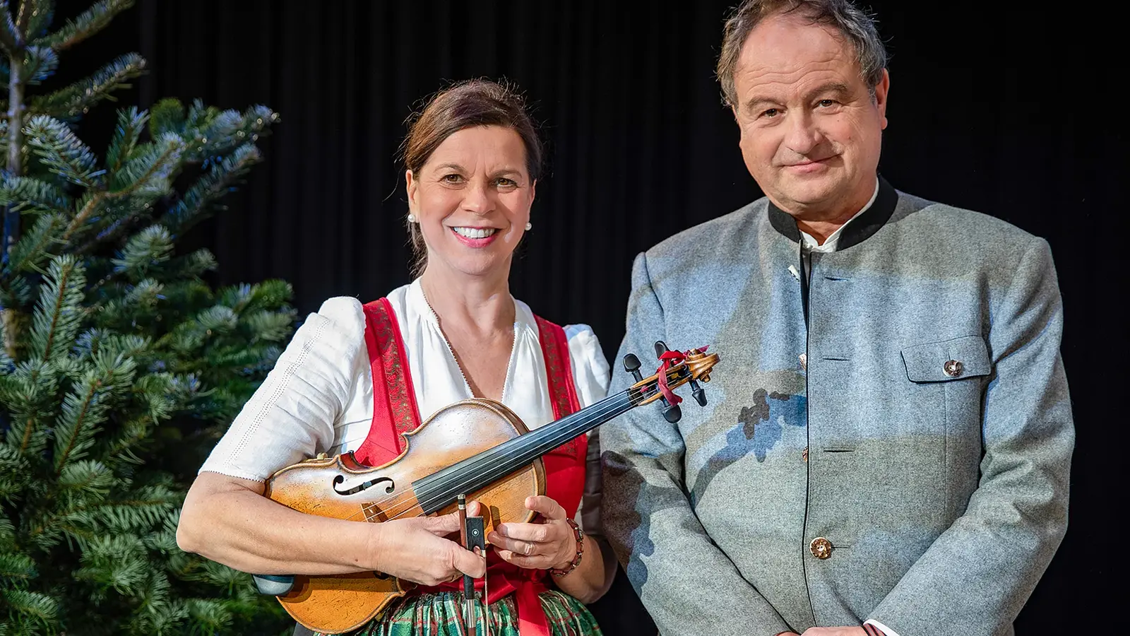 Zwei Personen neben einem Weihnachtsbaum vor schwarzem Hintergrund, eine Person in roter Tracht hält eine Violine in den Händen, die andere trägt eine graue Trachtenjacke