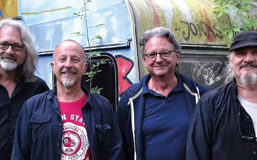 Vier Personen stehen nebeneinander vor einem alten, teilweise mit Graffiti besprühten Wohnwagen im Freien. Die Personen tragen überwiegend dunkle Freizeitkleidung, eine Person trägt ein rotes T-Shirt. Im Hintergrund sind Pflanzen und Bäume zu sehen, die dem Bild eine natürliche, rustikale Atmosphäre verleihen. Die Aufnahme zeigt die Gruppe in einer lockeren, informellen Umgebung.