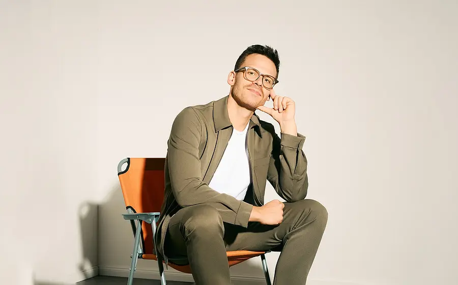 Martin Frank trägt ein grünes Outfit und sitzt vor einer hellen Wand auf einem orangenen Camping Stuhl.