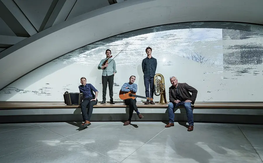 Copilot said:  Pressefoto der Band Maxjoseph mit fünf Musikern in moderner Architektur, darunter Violine, Akkordeon, Gitarre und Tuba, verteilt auf einer langen Sitzbank vor einer großen geschwungenen Fensterfront.