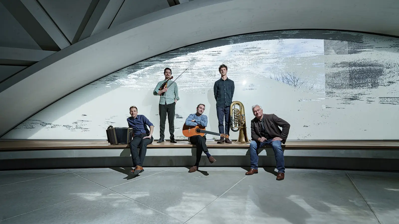 Copilot said: Pressefoto der Band Maxjoseph mit fünf Musikern in moderner Architektur, darunter Violine, Akkordeon, Gitarre und Tuba, verteilt auf einer langen Sitzbank vor einer großen geschwungenen Fensterfront.