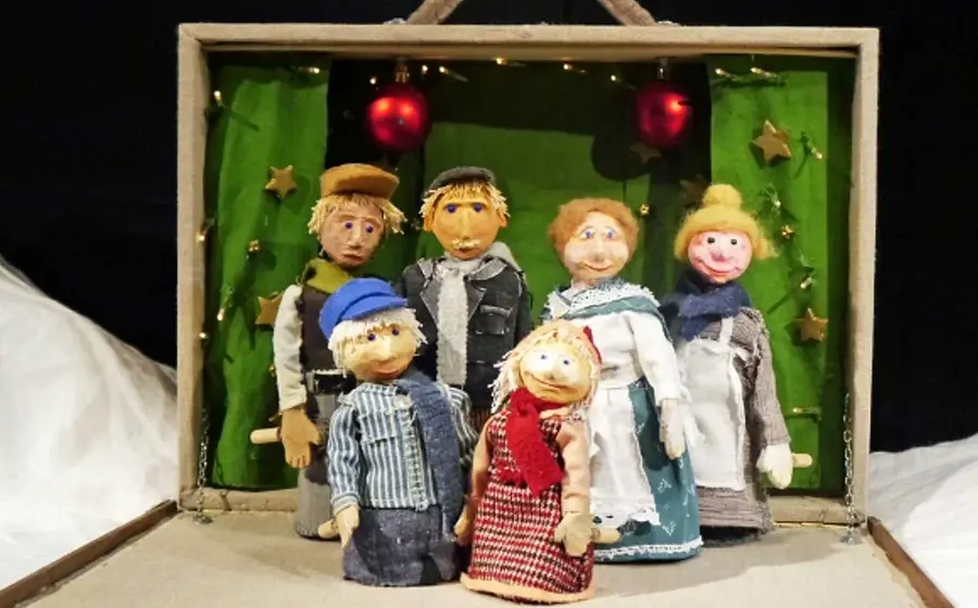 Bühnenbild aus Michel feiert Weihnachten mit sechs handgefertigten Figuren in Winterkleidung vor einer grünen Kulisse in einem geöffneten Puppentheaterkasten, dekoriert mit Sternen, Lichterkette und roten Christbaumkugeln.