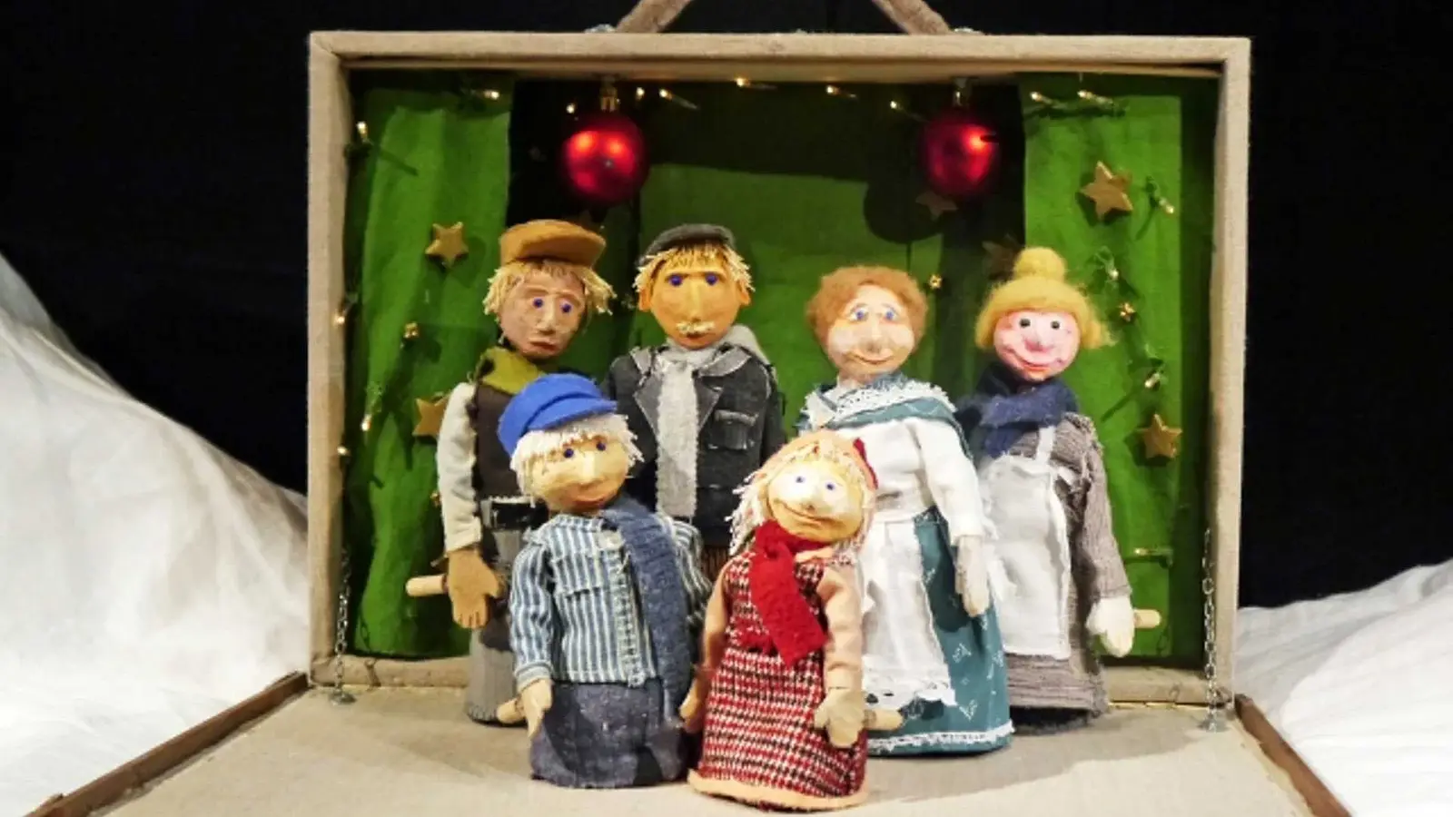 Bühnenbild aus Michel feiert Weihnachten mit sechs handgefertigten Figuren in Winterkleidung vor einer grünen Kulisse in einem geöffneten Puppentheaterkasten, dekoriert mit Sternen, Lichterkette und roten Christbaumkugeln.