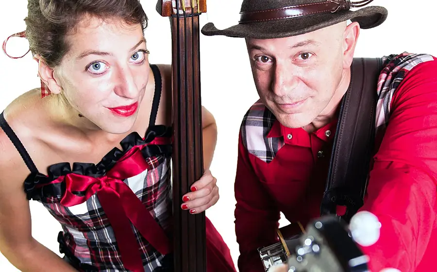 Zwei Personen im rot karierten Bühnenoutfit mit Kontrabass und Gitarre blicken in die Kamera