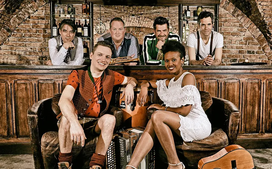 Fünf Musiker*innen der Gruppe CubaBoarisch sitzen in einer rustikalen Bar; vorne ein Mann in Tracht und eine Frau im weißen Kleid, dahinter drei Männer mit Trompete und ein weiterer Mann.
