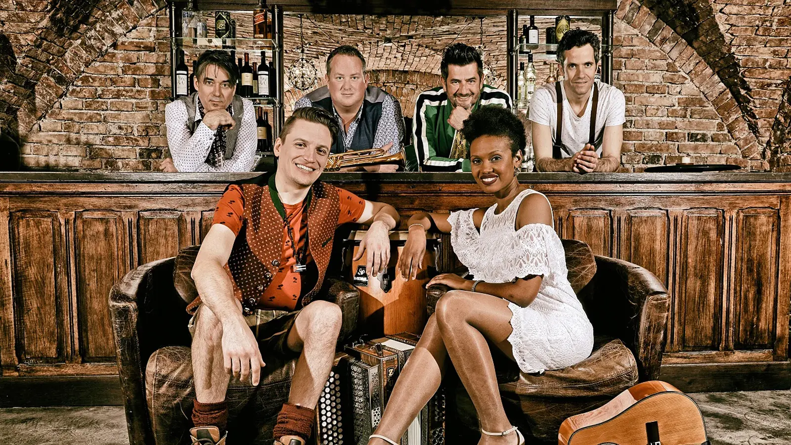 Fünf Musiker*innen der Gruppe CubaBoarisch sitzen in einer rustikalen Bar; vorne ein Mann in Tracht und eine Frau im weißen Kleid, dahinter drei Männer mit Trompete und ein weiterer Mann.