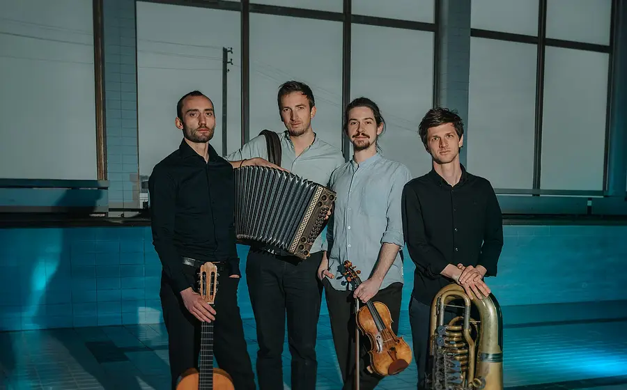Vier Musiker stehen nebeneinander in einem leeren Hallenbad. Sie halten Gitarre, Akkordeon, Violine und Tuba in den Händen. Große Fenster und das blau beleuchtete Becken bilden den Hintergrund.