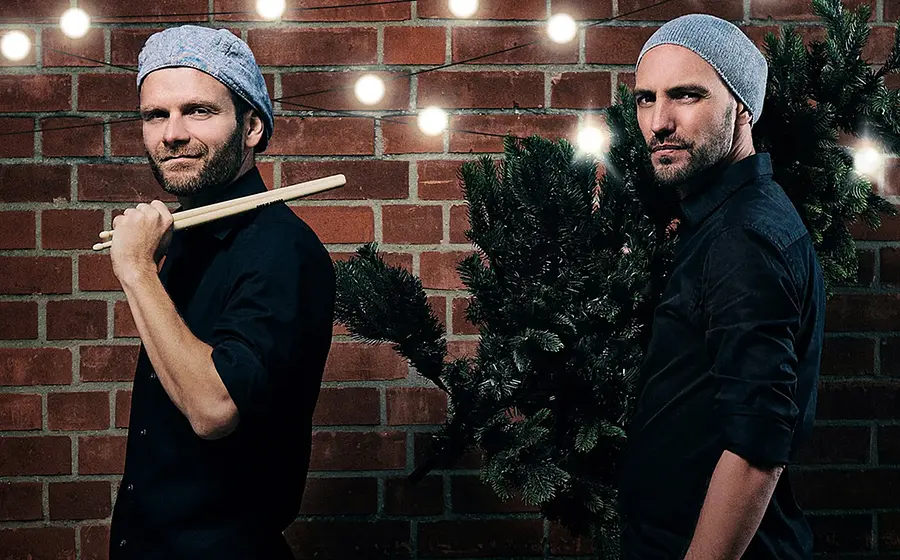 Pressefoto von Double Drums mit zwei Personen in schwarzen Hemden und grauen Mützen vor einer Backsteinwand, eine Person mit Drumsticks auf der Schulter, die andere mit einem geschmückten Tannenbaum auf dem Rücken, darüber Lichterkette mit warmen Glühbirnen.
