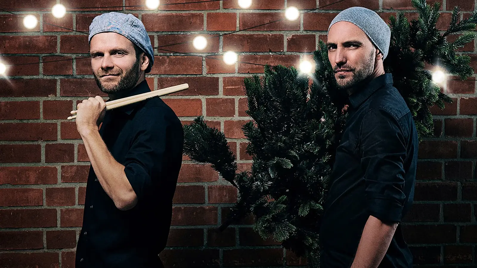 Pressefoto von Double Drums mit zwei Personen in schwarzen Hemden und grauen Mützen vor einer Backsteinwand, eine Person mit Drumsticks auf der Schulter, die andere mit einem geschmückten Tannenbaum auf dem Rücken, darüber Lichterkette mit warmen Glühbirnen.