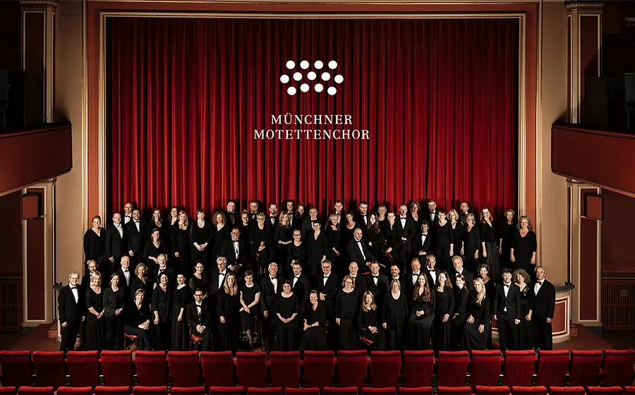 Große Chorgemeinschaft des Münchner Motettenchors in einem Theater mit roten Vorhängen und roten Sitzreihen. Die Sängerinnen und Sänger stehen auf der Bühne und im Zuschauerraum für ein Gruppenfoto.