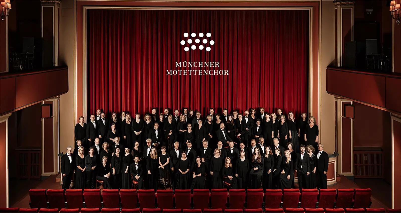 Große Chorgemeinschaft des Münchner Motettenchors in einem Theater mit roten Vorhängen und roten Sitzreihen. Die Sängerinnen und Sänger stehen auf der Bühne und im Zuschauerraum für ein Gruppenfoto.