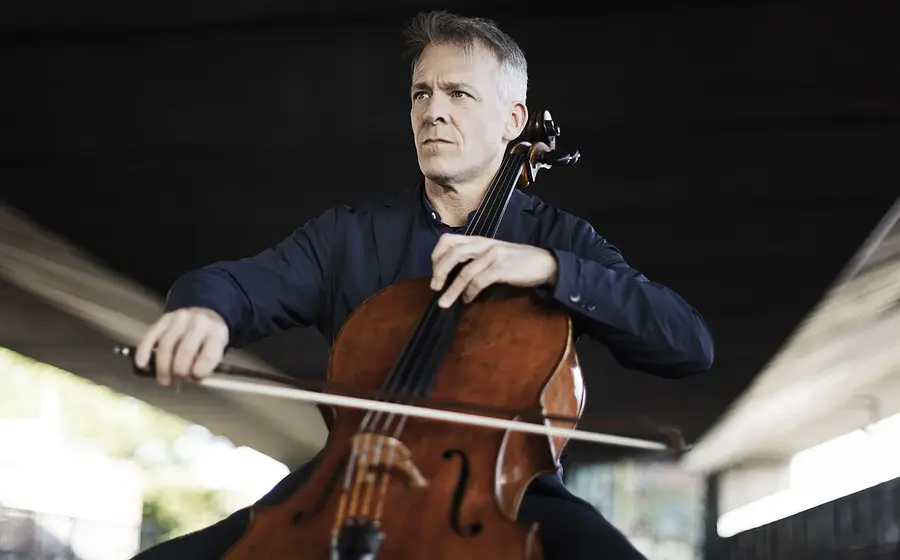 Eine Person in dunkelblauem Sakko, die Violoncello spielt, aufgenommen unter einer Brückenkonstruktion vor unscharfem Hintergrund