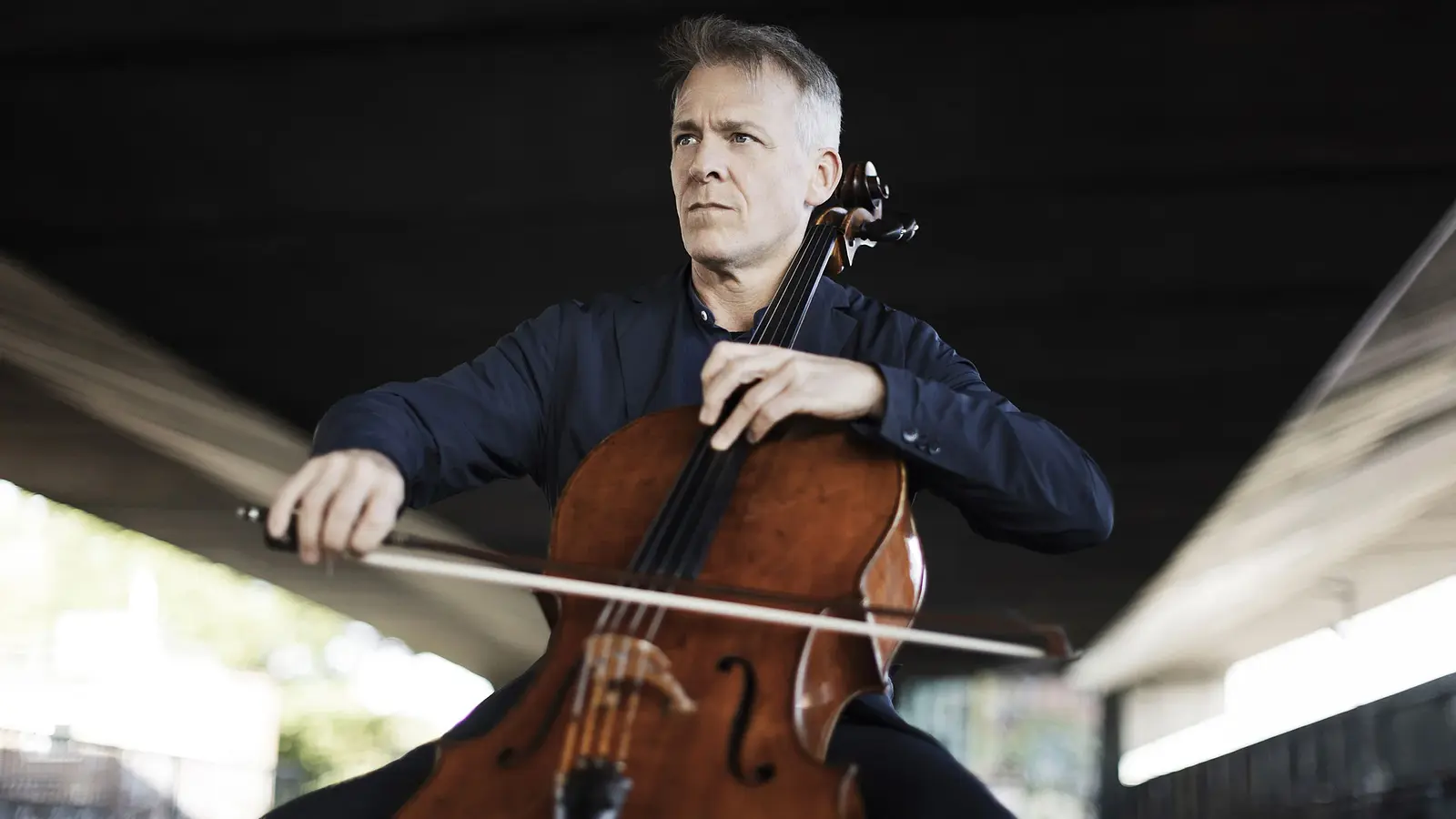 Eine Person in dunkelblauem Sakko, die Violoncello spielt, aufgenommen unter einer Brückenkonstruktion vor unscharfem Hintergrund