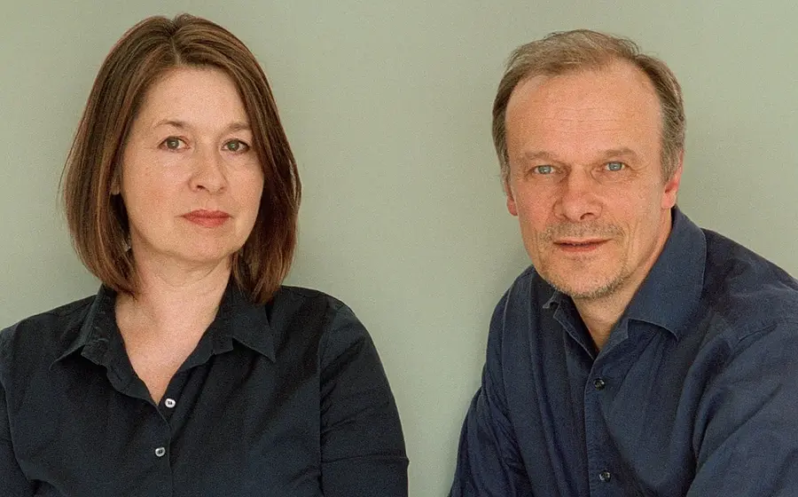 Edgar Selge und Franziska Walser in dunkler Kleidung sitzen nebeneinander vor neutralem Hintergrund und blicken in die Kamera.