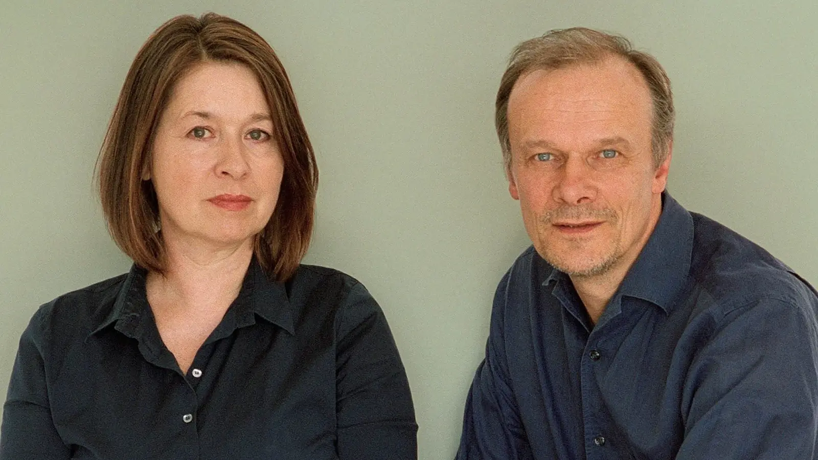 Edgar Selge und Franziska Walser in dunkler Kleidung sitzen nebeneinander vor neutralem Hintergrund und blicken in die Kamera.