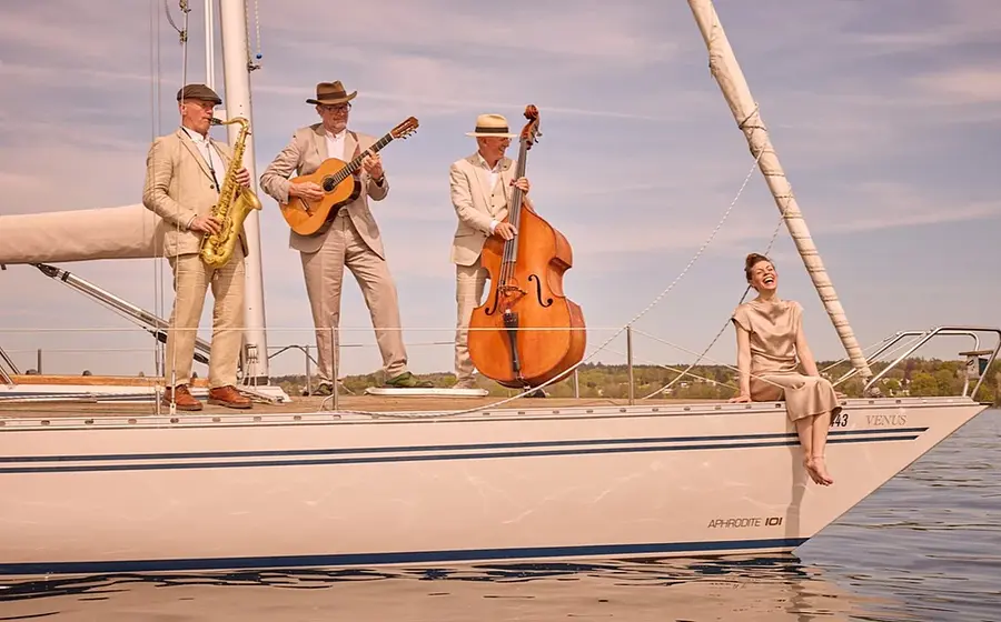 Pressefoto des Trio Nautico mit einer Sängerin und drei Musikern auf einer Segelyacht, darunter Saxofon, Gitarre und Kontrabass, auf einem See vor Uferlandschaft und hellem Himmel.