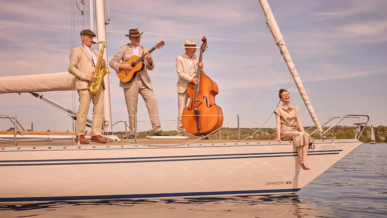 Pressefoto des Trio Nautico mit einer Sängerin und drei Musikern auf einer Segelyacht, darunter Saxofon, Gitarre und Kontrabass, auf einem See vor Uferlandschaft und hellem Himmel.