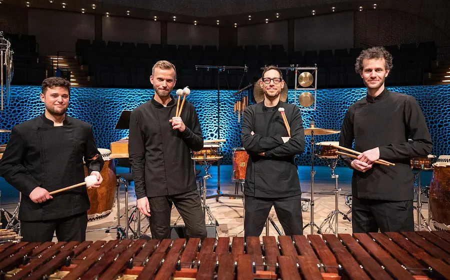 Vier Percussionisten stehen zwischen verschiedenen Schlaginstrumenten auf einer Konzertbühne und halten Schlägel in den Händen