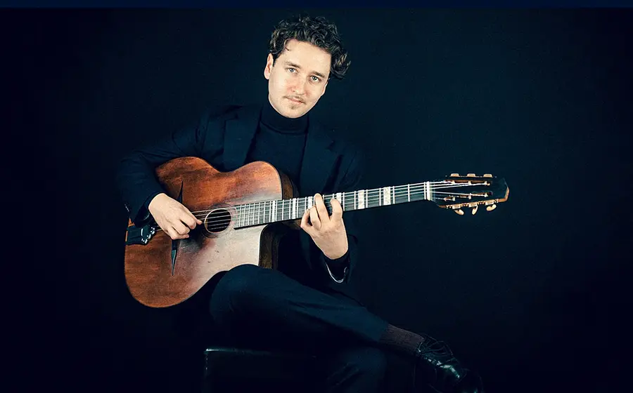 Porträtfoto von Elias Prinz mit Akustikgitarre, sitzend auf einem Hocker vor schwarzem Hintergrund, in dunklem Anzug und Rollkragenpullover während des Gitarrenspiels